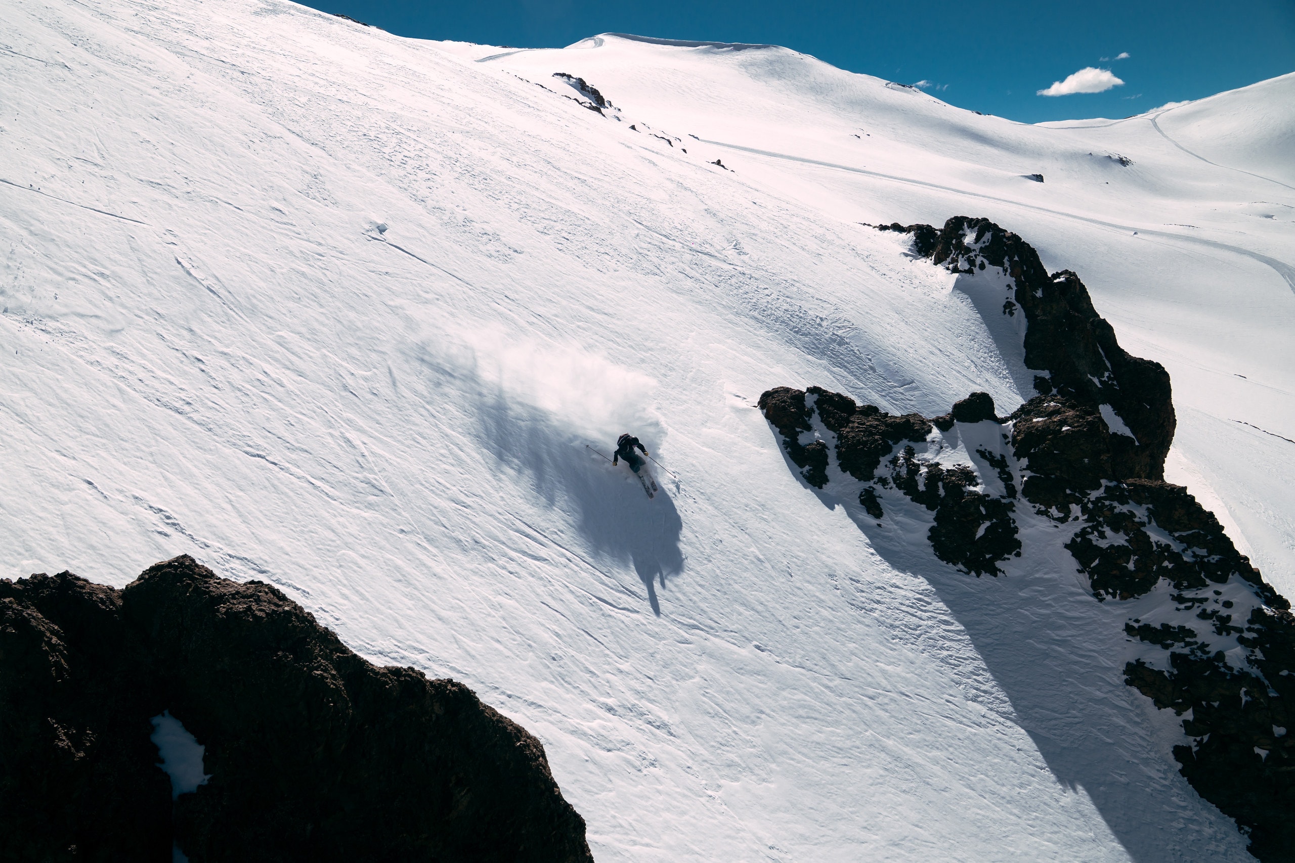 A skier carving down a wide snowfield between bands of dark rock, spray hanging behind