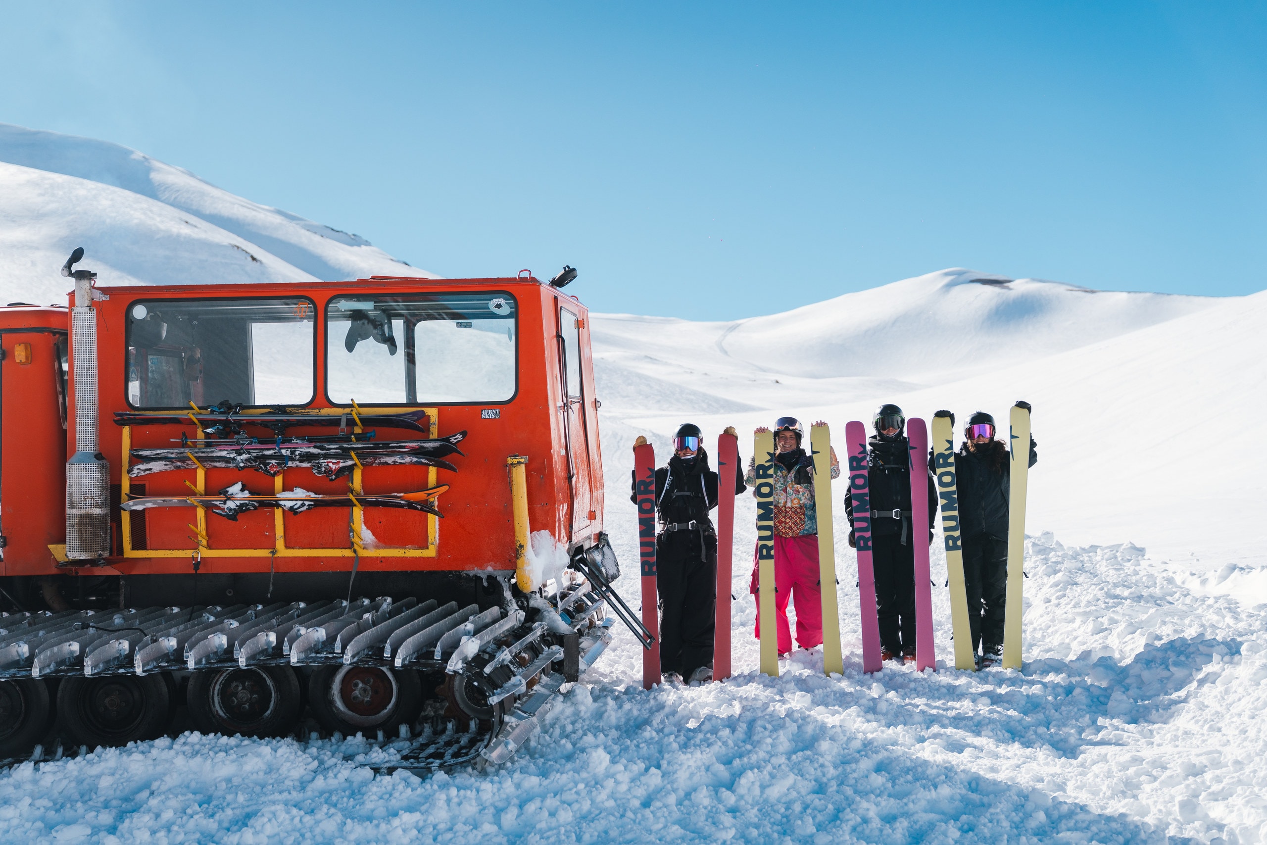 The RUMOR crew lined up beside an orange snowcat, holding their skis upright